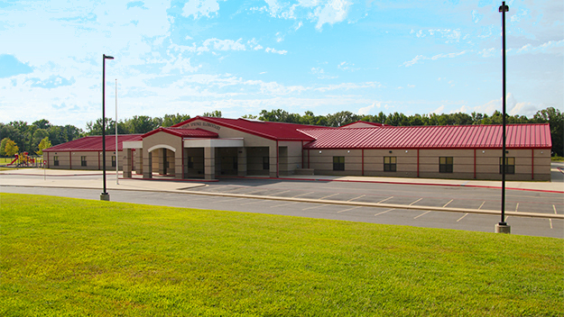 A single-story school building with a red roof and beige walls, surrounded by green grass and an empty parking lot, with trees and a partly cloudy sky in the background.