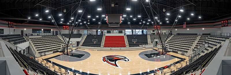 Wide-angle view of an empty indoor basketball arena with tiered seating, a wooden court featuring a large hawk logo at center, and electronic scoreboards above each basket.