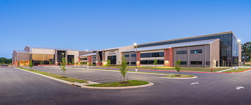 Modern school building with large windows and brick accents, surrounded by an empty parking lot and small trees, under a clear blue sky at dusk.