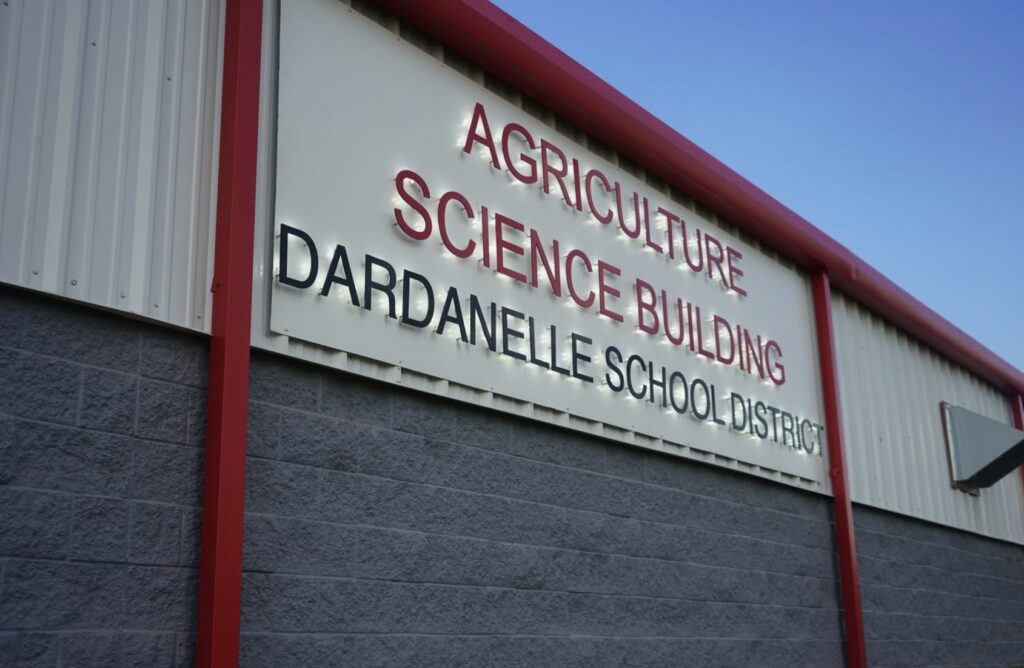 A building with a sign that reads Agriculture Science Building Dardanelle School District against a clear blue sky. The sign has red and black lettering on a white background.