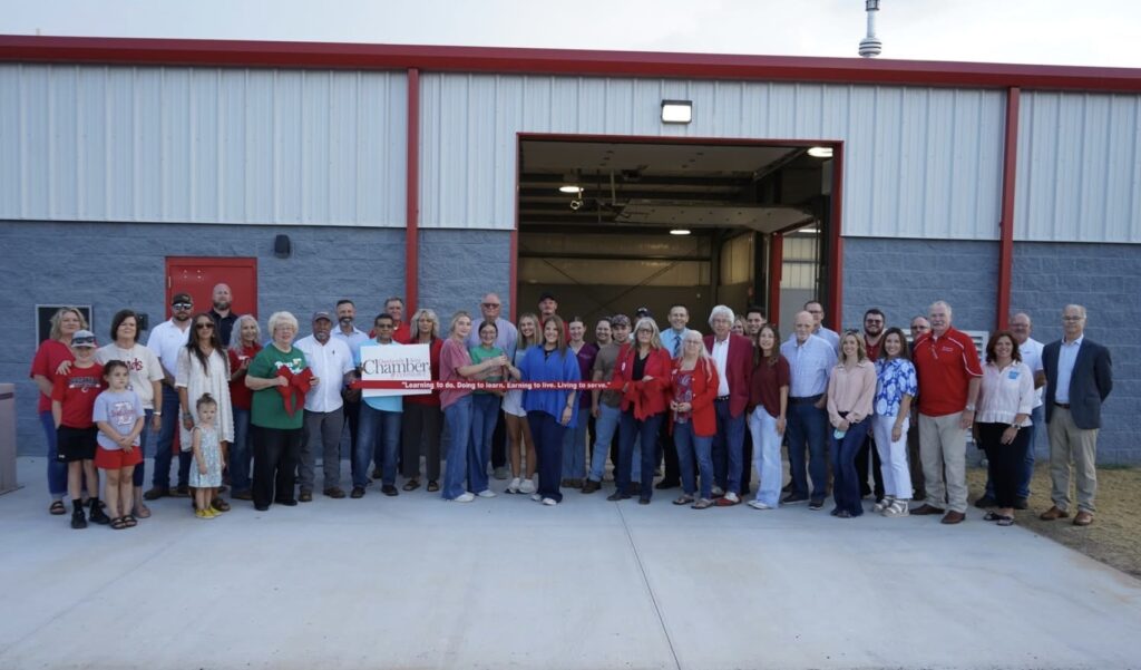 A large group of people of various ages poses in front of a gray and red metal building, holding a ribbon for a ribbon-cutting ceremony. The building’s large garage door is open behind them.