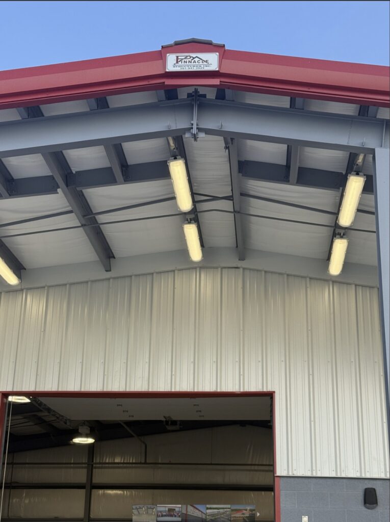 A large metal garage with red trim and an open entrance, showing fluorescent lights on the high ceiling and a Princeton Construction sign above the doorway.