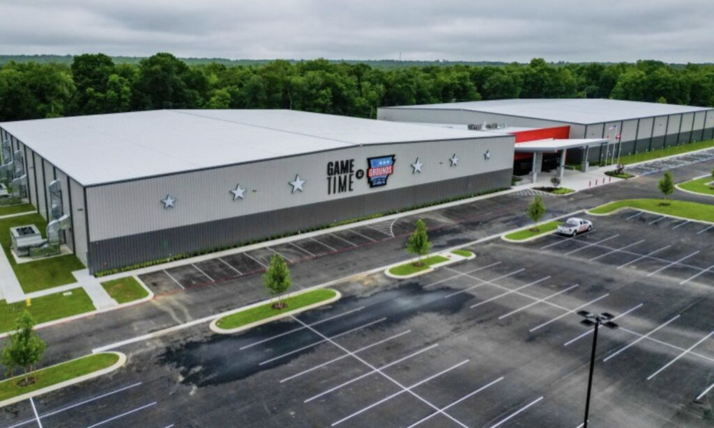 A large sports complex with Game Time and Athletic Training & Fitness signs on the building front, surrounded by an empty parking lot and green trees under a cloudy sky.