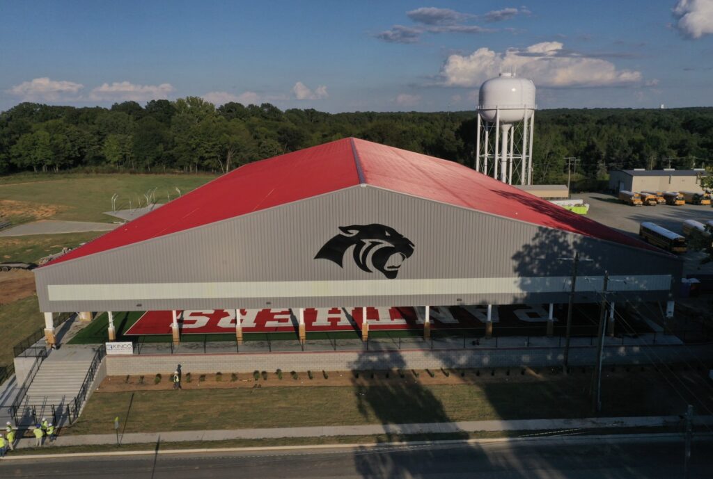 A large sports facility with a red roof and a panther logo on the side stands next to a football field marked PANTHERS. A white water tower and trees are visible in the background under a blue sky.