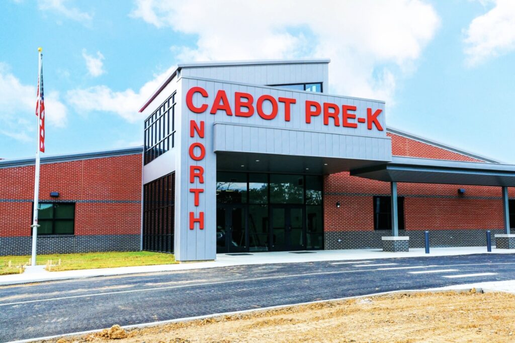 A modern brick and metal school building with large red letters reading CABOT PRE-K NORTH above the entrance, under a partly cloudy sky. A newly paved road and some dirt are visible in front.