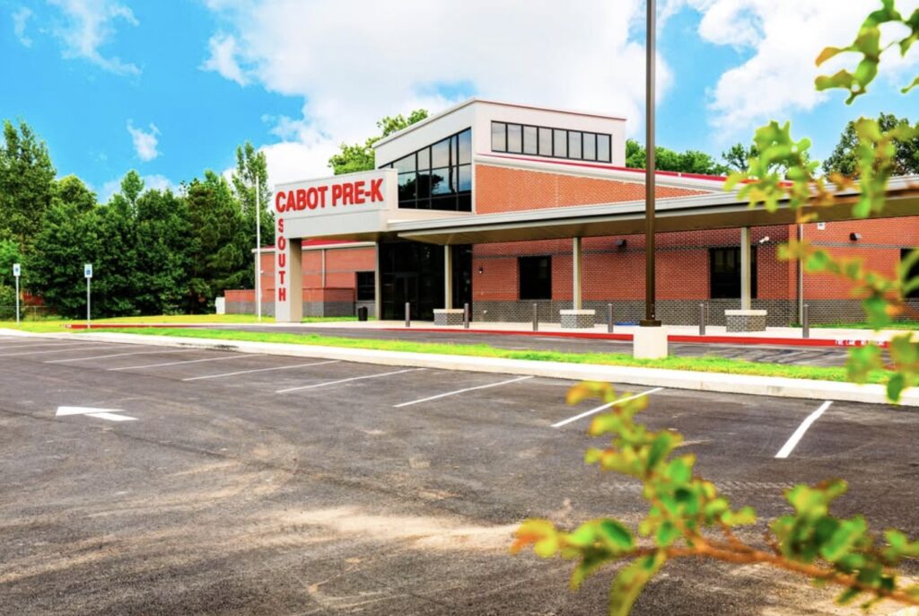 A modern, brick school building with large windows and a sign reading Cabot Pre-K above the entrance. The parking lot in front is empty, and leafy green trees surround the building under a bright blue sky.