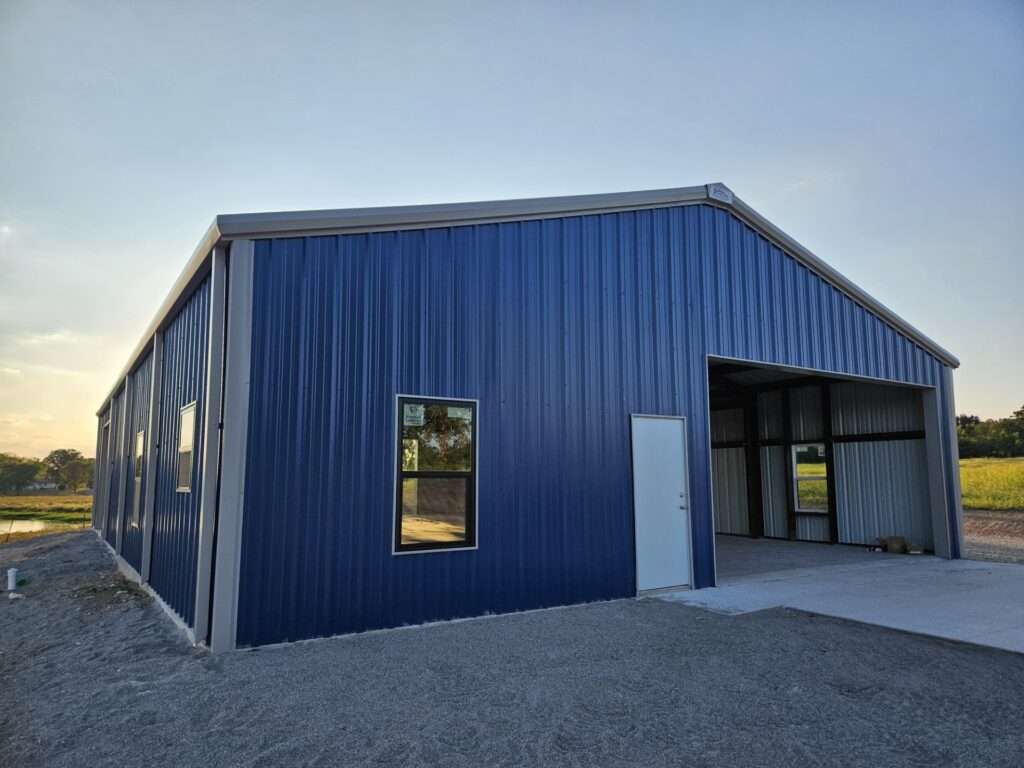 A large blue metal building with a sloped roof, several windows, a white door, and an open garage space, set on a gravel surface with a grassy field in the background under a clear sky.