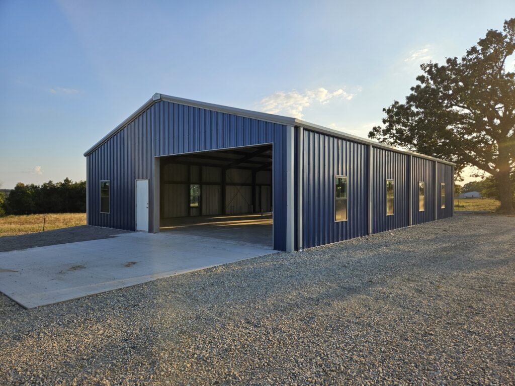 A large blue metal building with an open garage door, concrete driveway, and several windows stands on a gravel lot under a clear sky in a rural setting near a tree.
