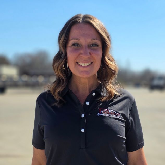 A woman with wavy, shoulder-length brown hair smiles at the camera. She is wearing a black collared shirt with an embroidered logo and is standing outdoors on a sunny day.
