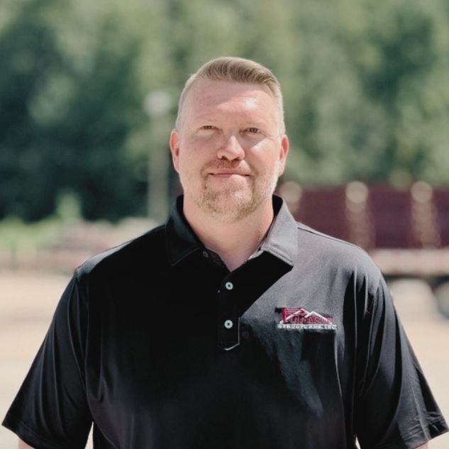 A man with short blond hair and a light beard stands outdoors, wearing a black polo shirt with a red and white embroidered logo, with trees blurred in the background.