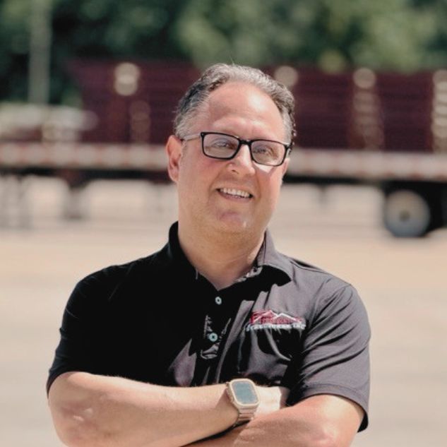 A man with glasses, wearing a black polo shirt and smartwatch, stands outdoors with arms crossed, smiling. A blurred truck and trees are in the background.